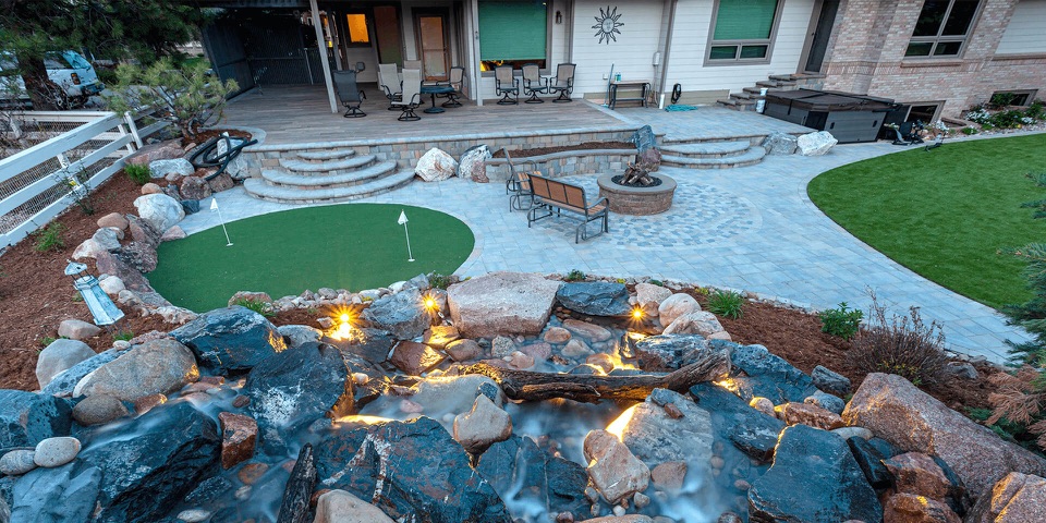 Backyard at twilight: putting green, patio, boulders, lit waterfall