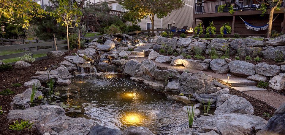 A natural looking landscape waterfall in backyard with pond next to stone stairs and gravel walkway
