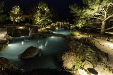 Natural swimming pond lit at night with a grotto waterfall and pergola, Peyton