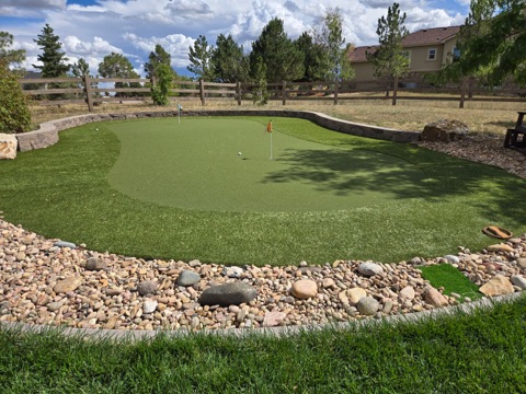 Backyard artificial turf putting green with two flags, framed by a curved stone retaining wall and a river rock border in Colorado