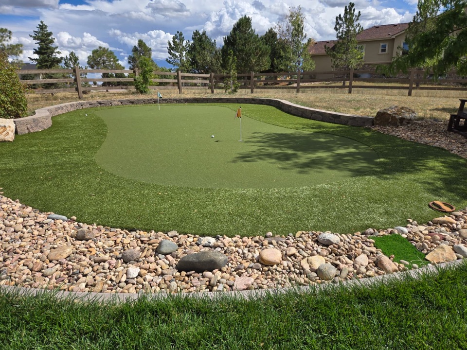 Backyard artificial turf putting green with two flags, framed by a curved stone retaining wall and a river rock border in Colorado