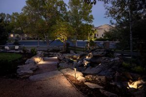 a backyard at dusk lit with lawn lights and a gravel walkway leading to a seating area in the distance