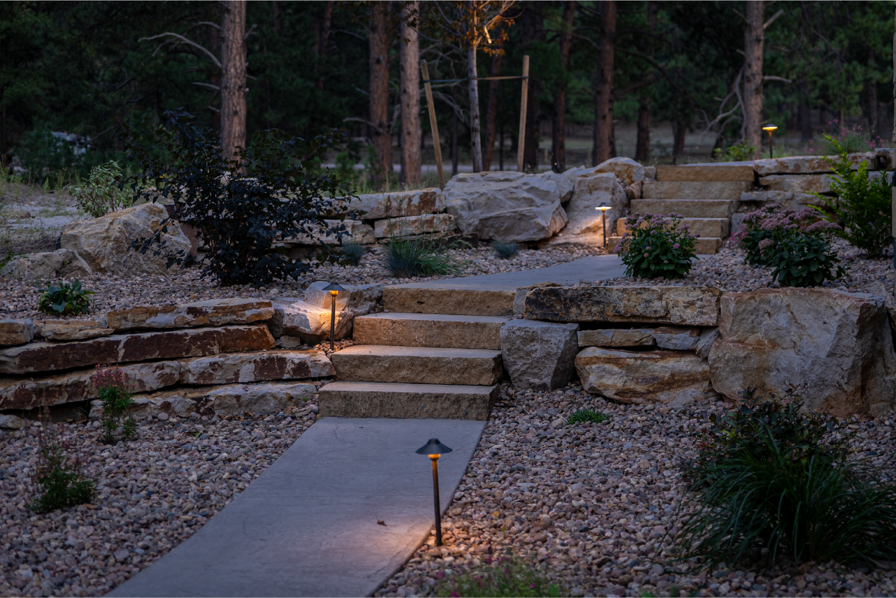 A stone staircase sits in the center of a drought-friendly landscape design with a walkway, lighting, gravel, and native plant life